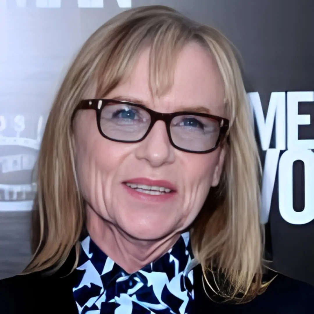 Woman with blonde hair and glasses, wearing a patterned blouse, smiling at the camera.