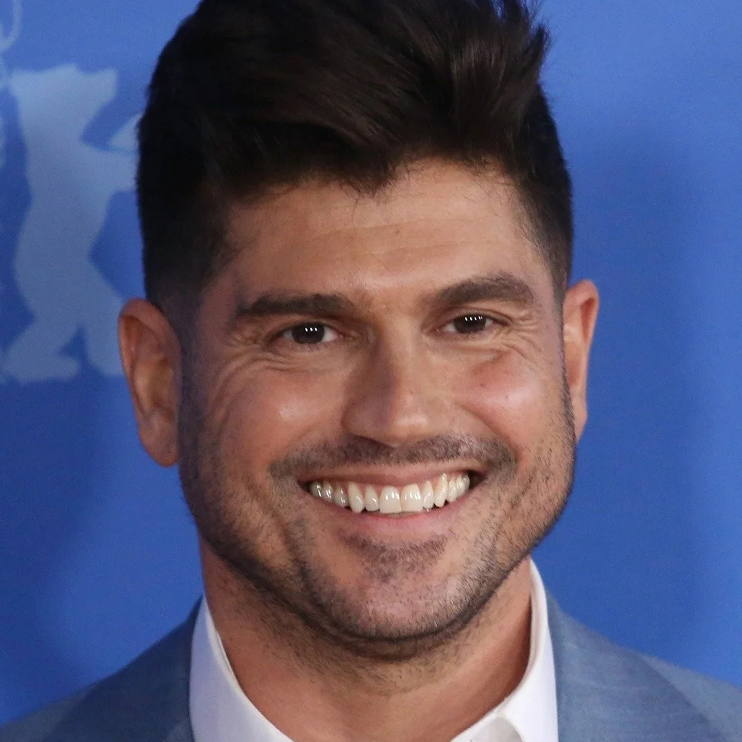 German actor Misel Maticevic smiling at a red carpet event, wearing a blue suit and white shirt.