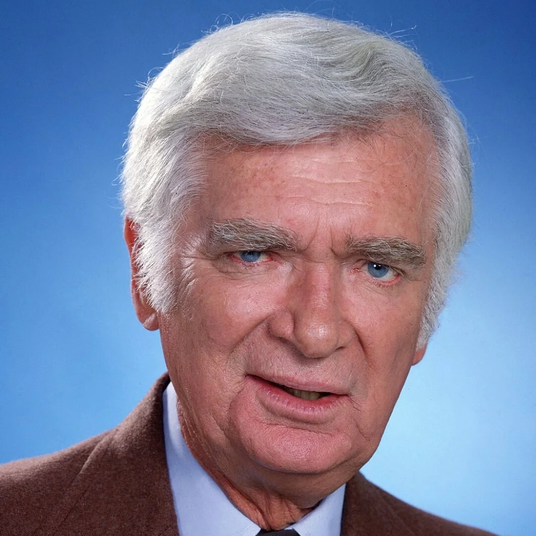 Portrait of an older man with white hair and blue eyes, wearing a brown suit against a blue background.