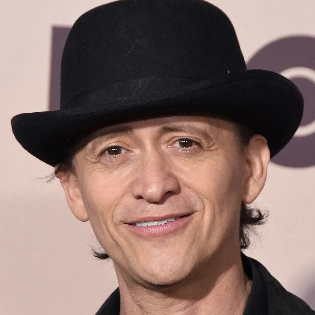 Portrait of a man wearing a black hat and a black jacket, smiling at the camera against a light-colored background.