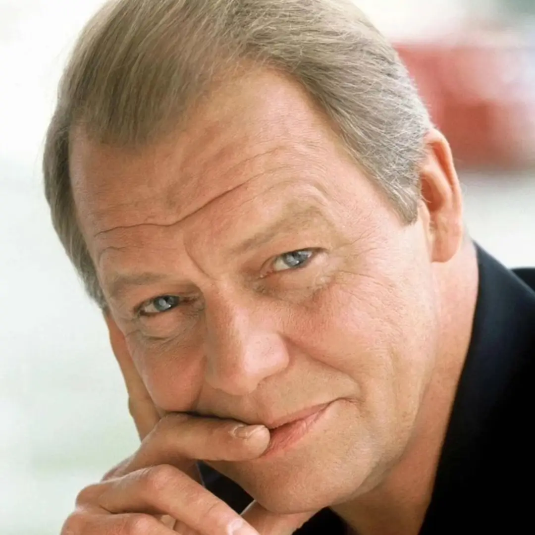 Close-up of an older man with blue eyes and light hair, resting his chin on his hand.