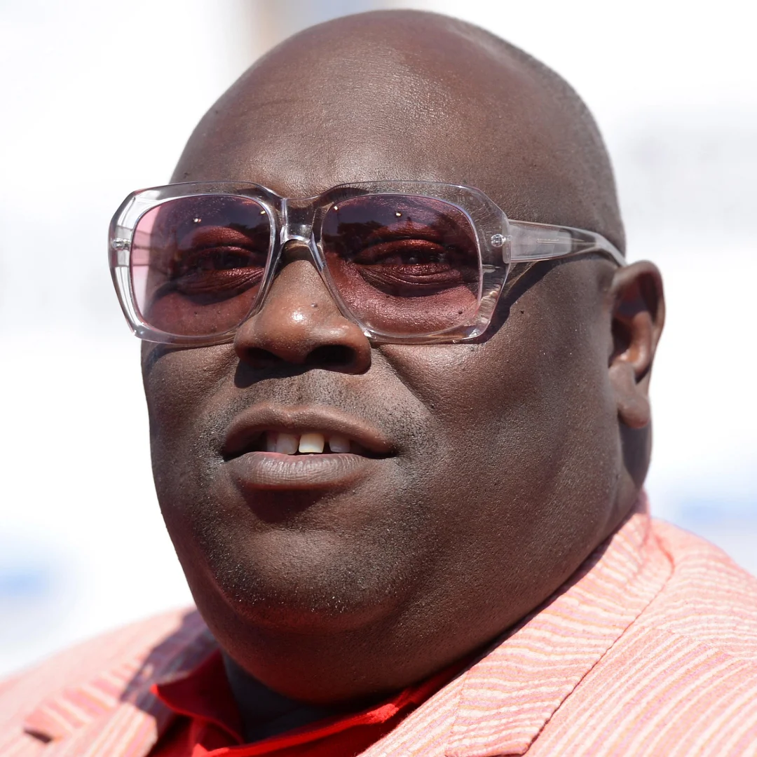 Close-up of a man with dark skin wearing transparent glasses and a light pink jacket.