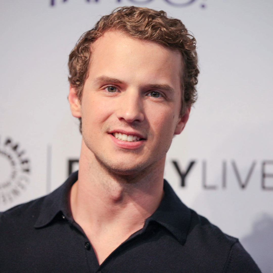 Portrait of a man with short curly hair, wearing a dark shirt, smiling at the camera, standing against a backdrop with text.