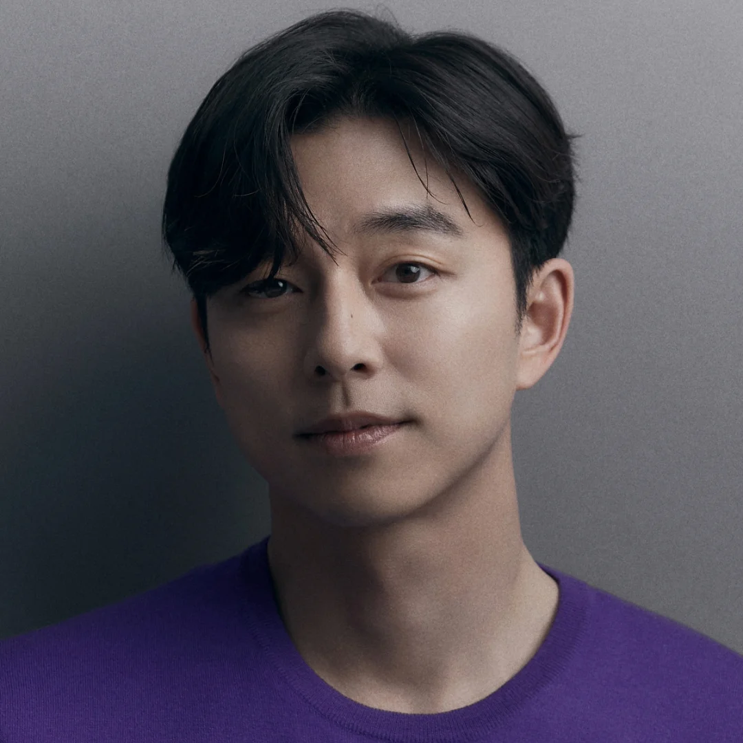 Portrait of a man with short dark hair, wearing a purple shirt, standing against a plain gray background.