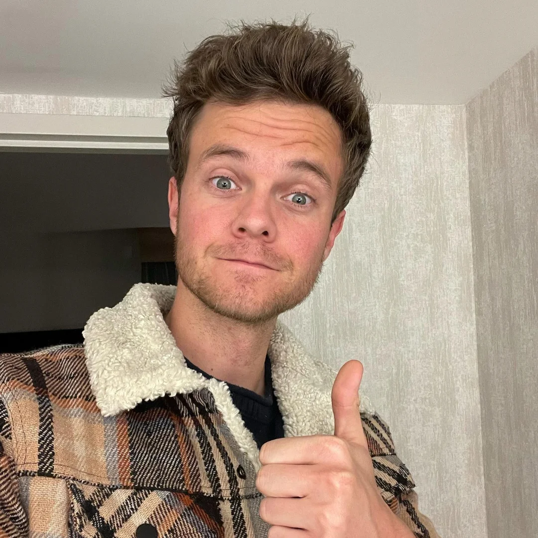 A young man with tousled hair giving a thumbs up while wearing a plaid jacket with a shearling collar.