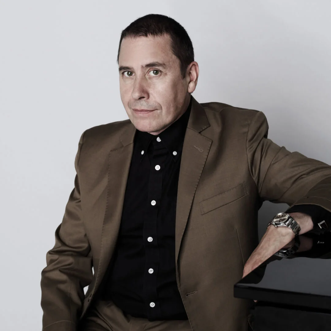 Man in a brown suit and black shirt, sitting with one arm resting on a surface.