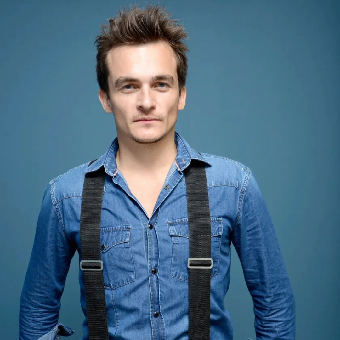 Rupert Friend in a denim shirt with suspenders, standing against a blue background.