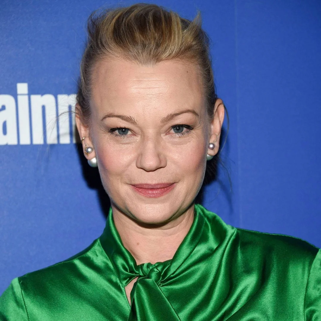 Portrait of a woman with blonde hair styled back, wearing a green satin blouse, standing against a blue background with text.