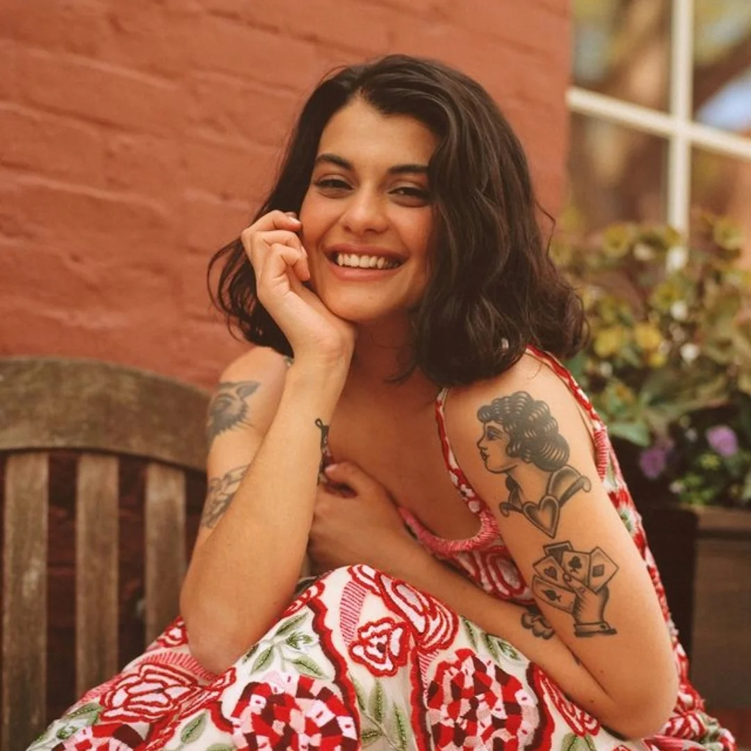 Smiling woman with tattoos, wearing a patterned dress, sitting in front of a brick wall.