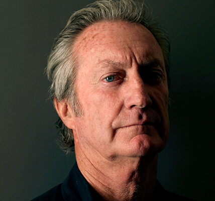 An older man with gray hair, wearing a dark shirt, is looking at the camera with a neutral expression.