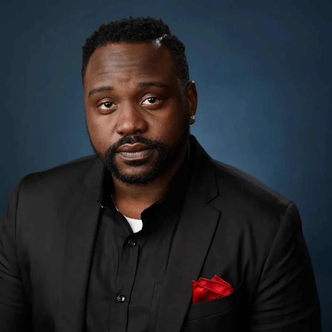 Brian Tyree Henry posing in a black suit with a red pocket square against a blue background