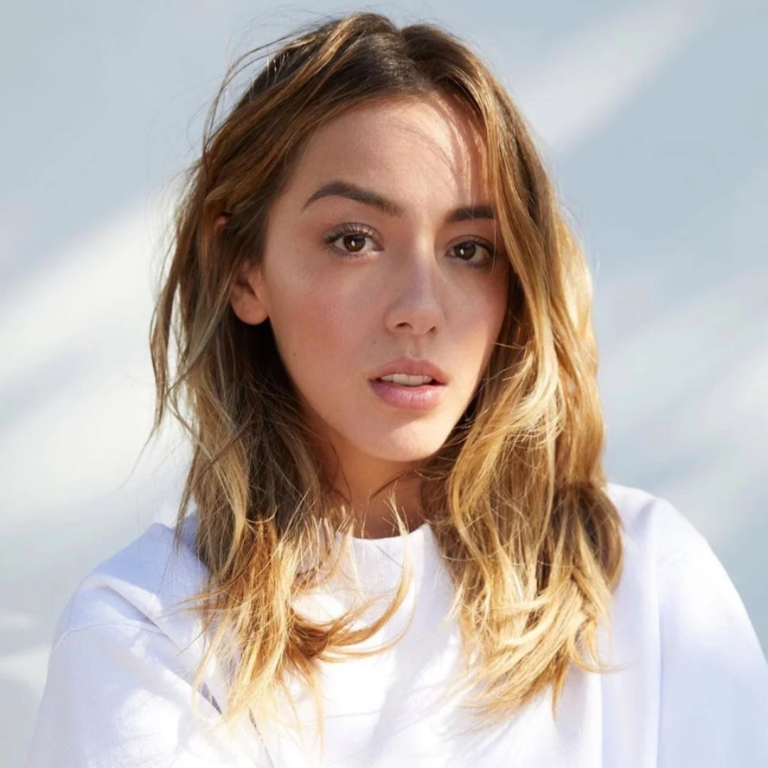 Woman with wavy blonde hair and natural makeup, wearing a white top, looking directly at the camera with a neutral expression, against a light background.