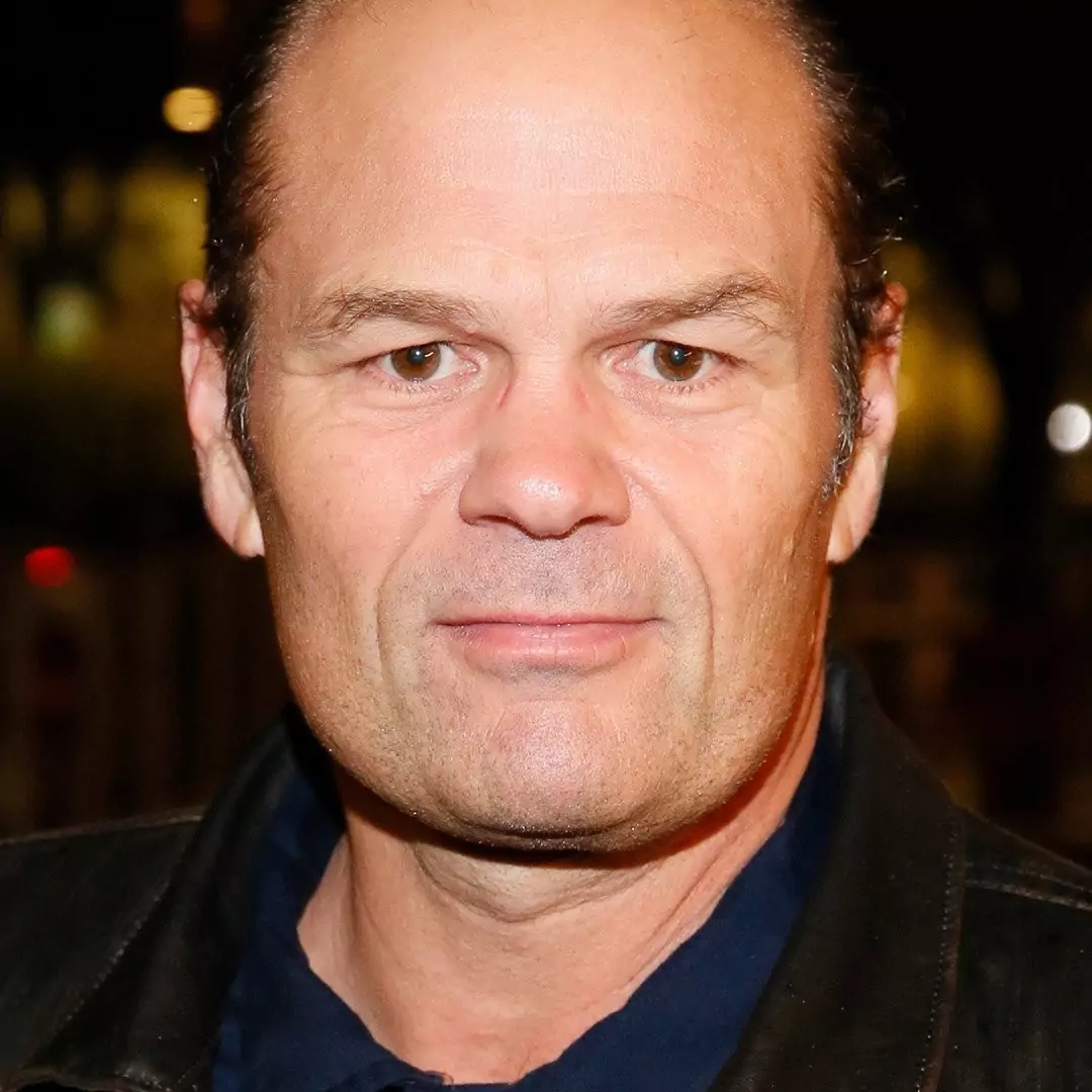 Portrait of Chris Bauer wearing a dark jacket and a navy blue shirt, standing outdoors at night.