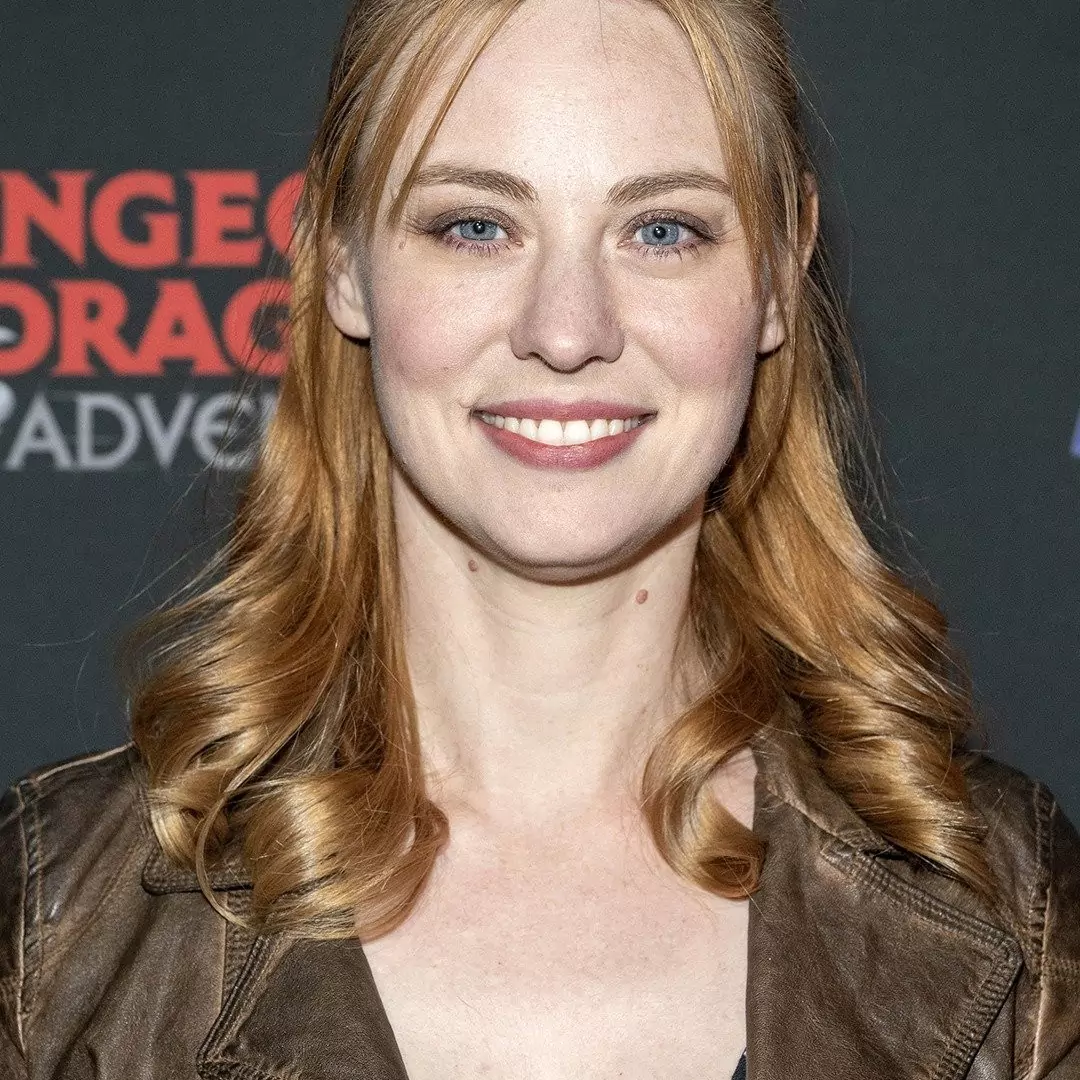 Deborah Ann Woll at a Dungeons & Dragons event, smiling and wearing a brown leather jacket with her red hair styled in soft curls