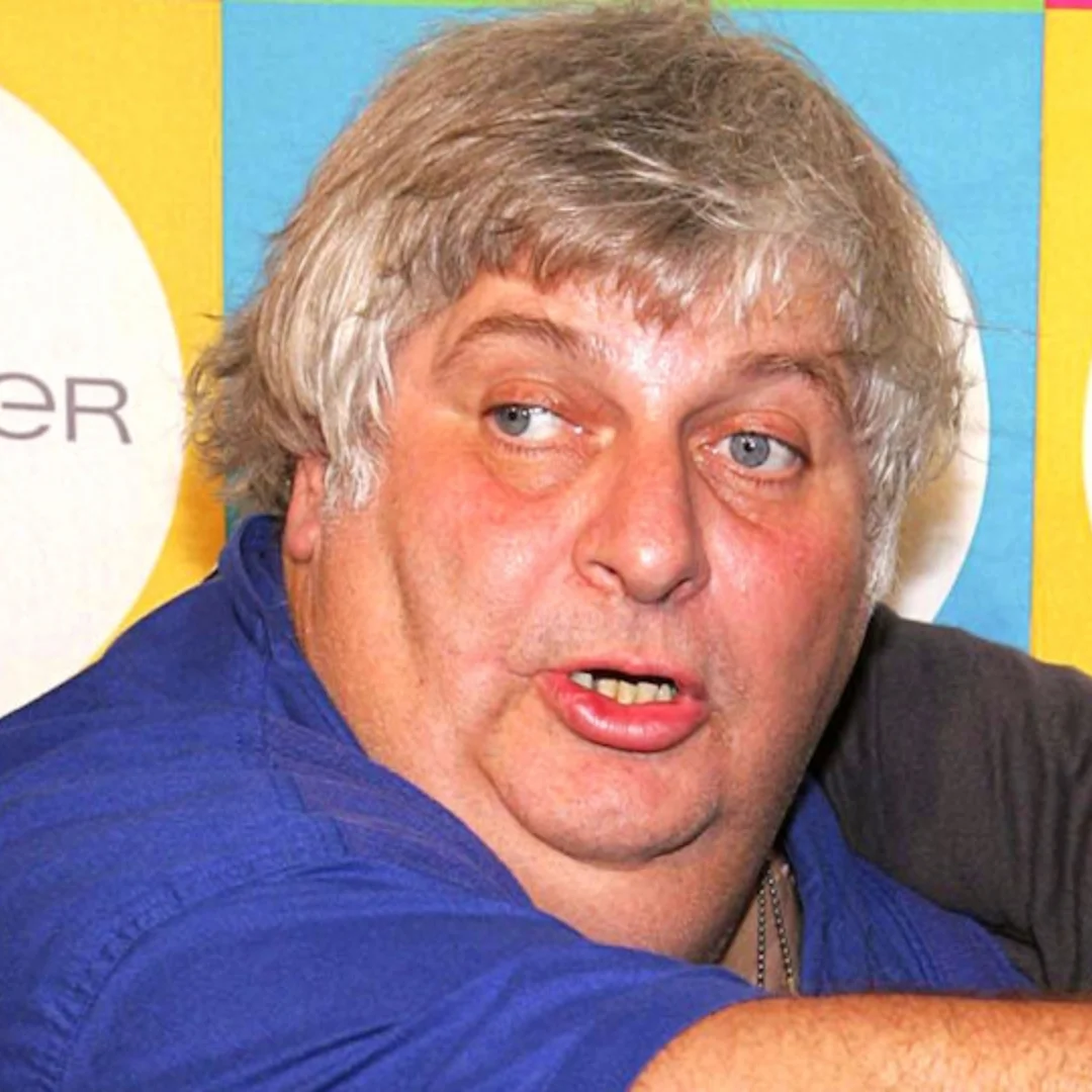 Don Vito Margera, a man with gray hair and blue eyes, in a blue shirt, making an expressive face at an event with colorful background