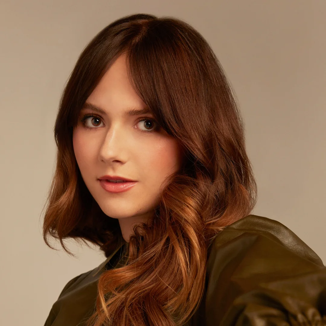 Young woman with long, wavy brown hair, looking directly at the camera with a subtle smile, wearing a dark green top, against a plain beige background.