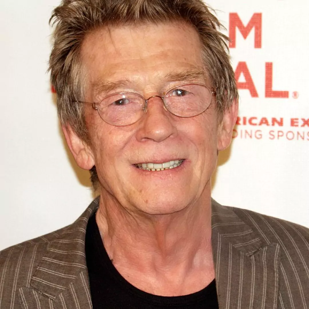 John Hurt at an event, wearing glasses and a striped suit, smiling with a neutral background