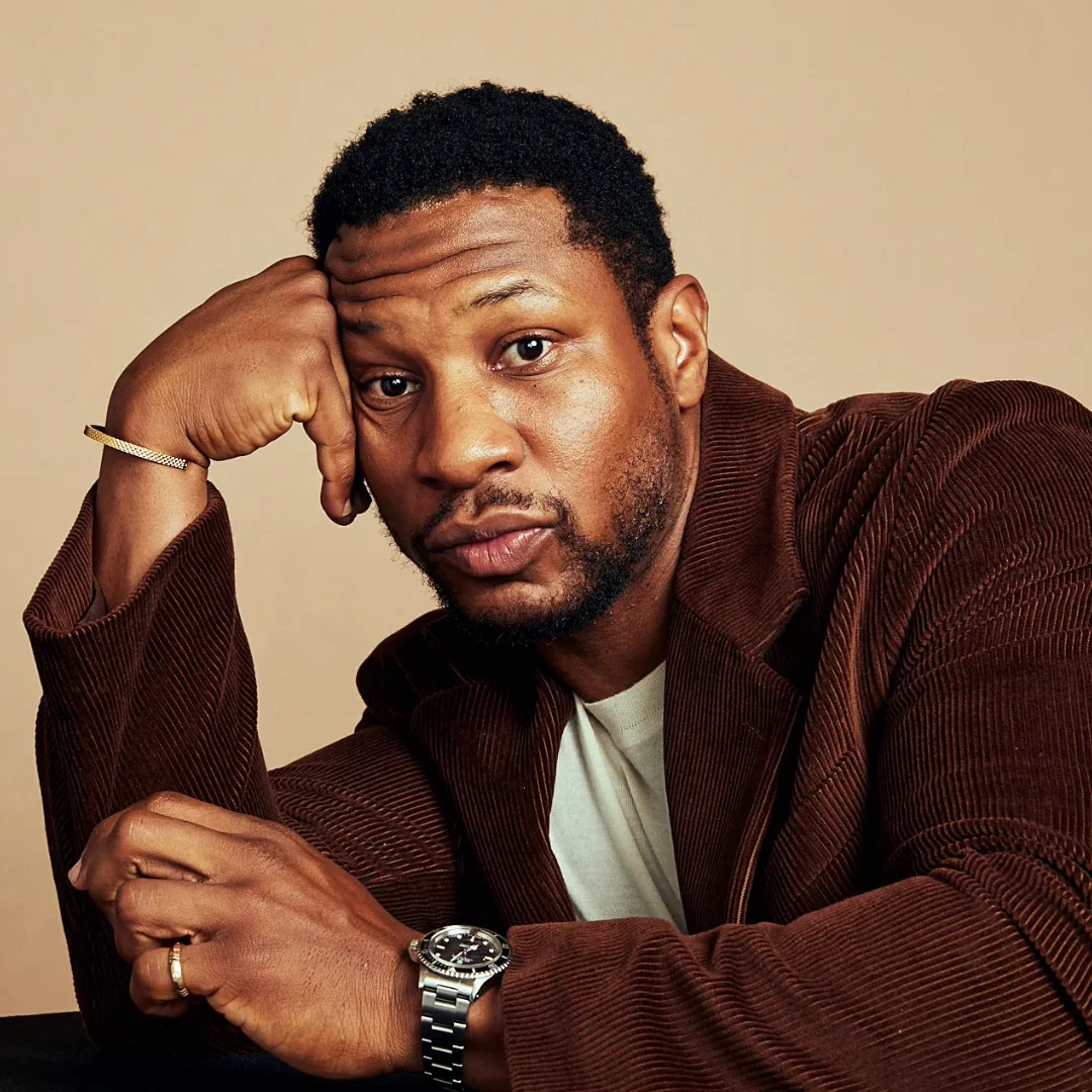 Jonathan Majors, American actor, posing in a thoughtful manner while wearing a brown jacket and watch.