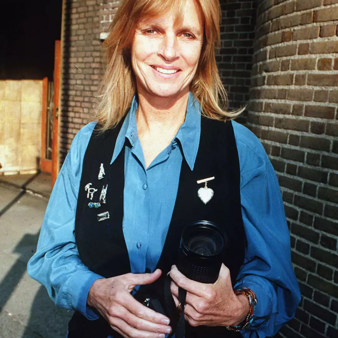 Linda McCartney holding a camera, wearing a blue shirt and black vest.