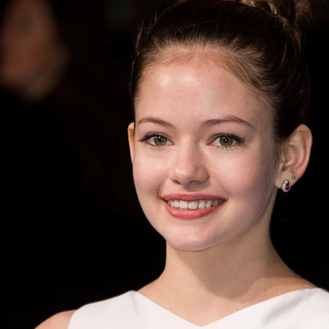 Young woman smiling, with her hair up, wearing earrings and a white top, against a dark background.