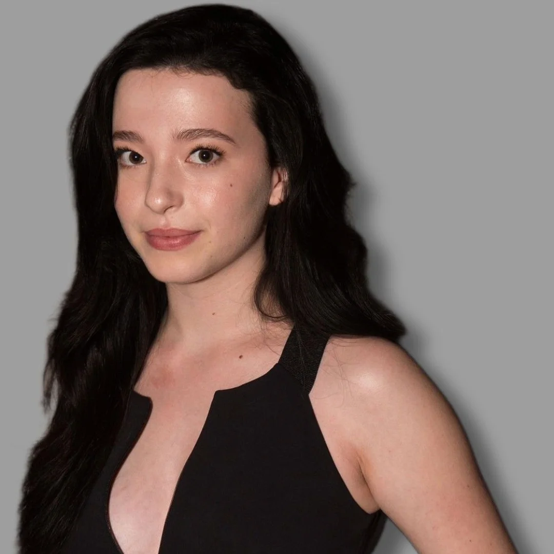 Woman with long dark hair, wearing a black sleeveless dress, smiling slightly against a gray background.