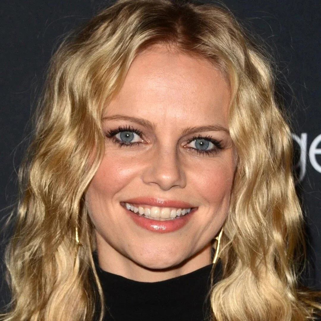 Close-up of a woman with wavy blonde hair, blue eyes, and a bright smile, wearing a black top and gold earrings, against a dark background