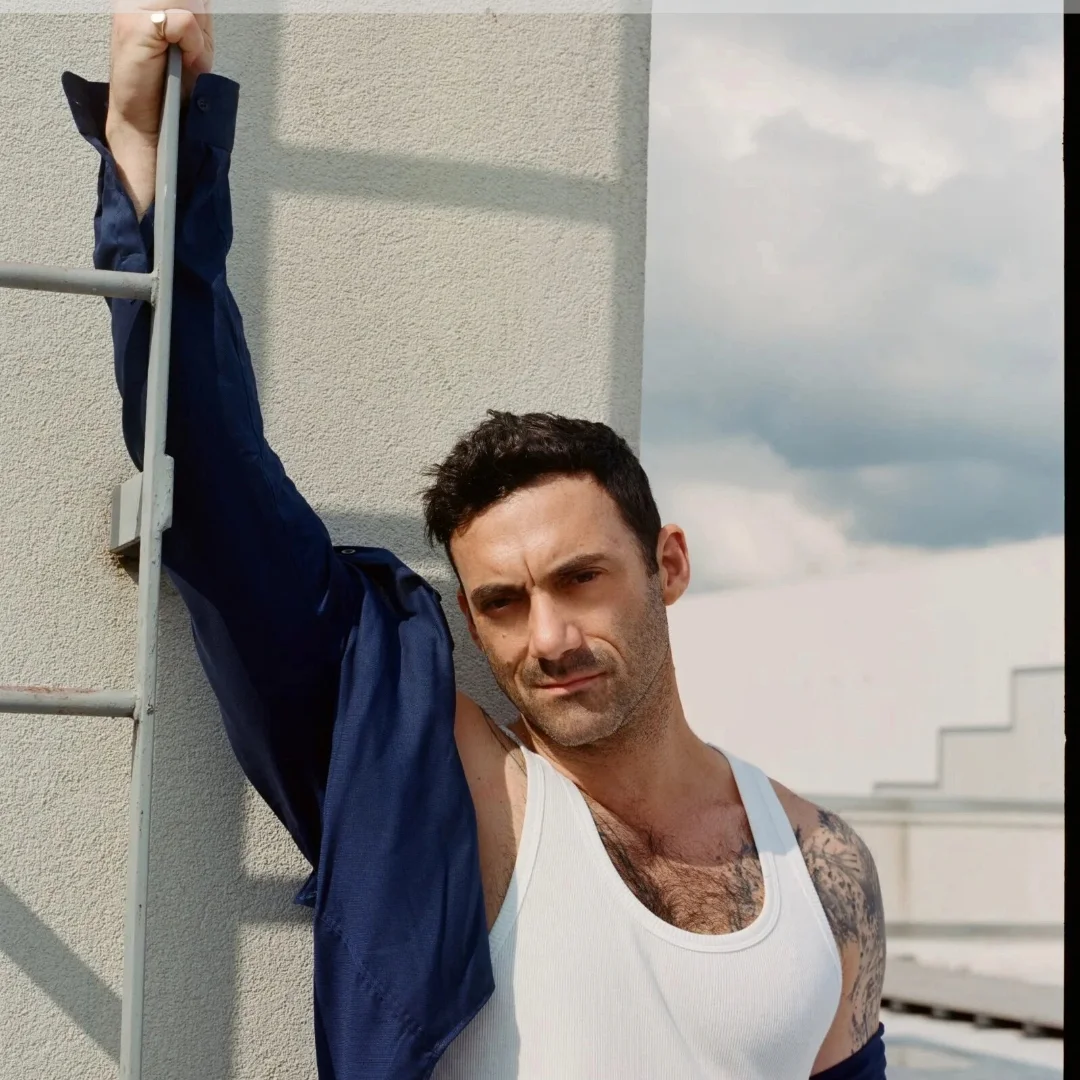 Man in a white tank top and blue jacket posing with one arm resting on a metal railing, standing against a concrete wall with a cloudy sky in the background.