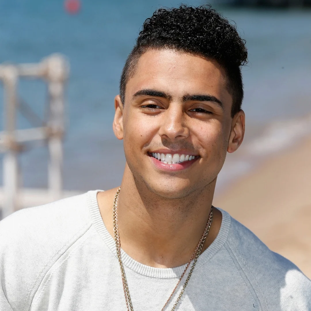 Man with short curly hair, wearing a light gray sweater, smiling at the camera with a beach and blue sea in the background