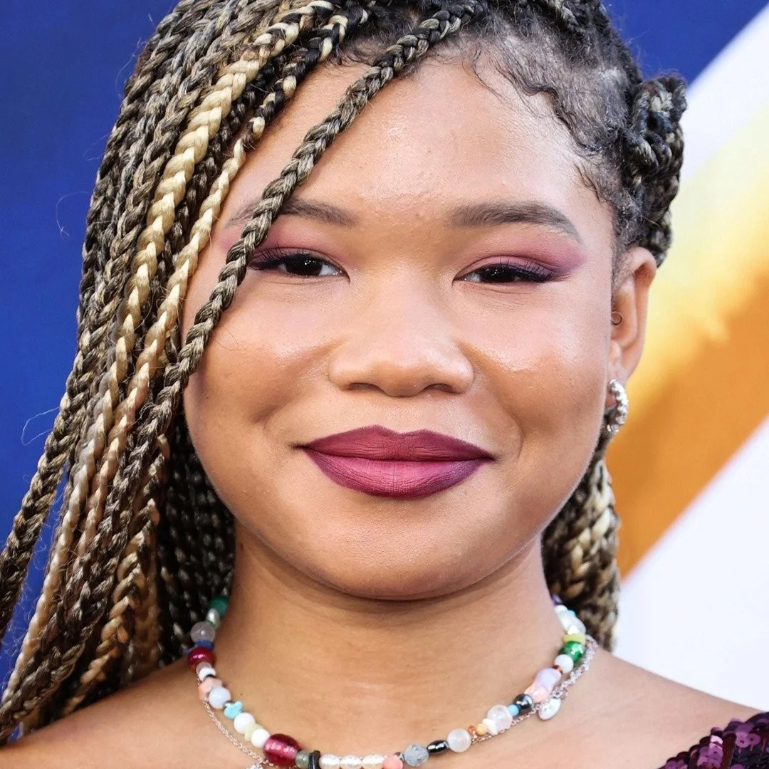 Woman with braided hair, wearing a colorful beaded necklace, and makeup with pink eyeshadow and dark lipstick.