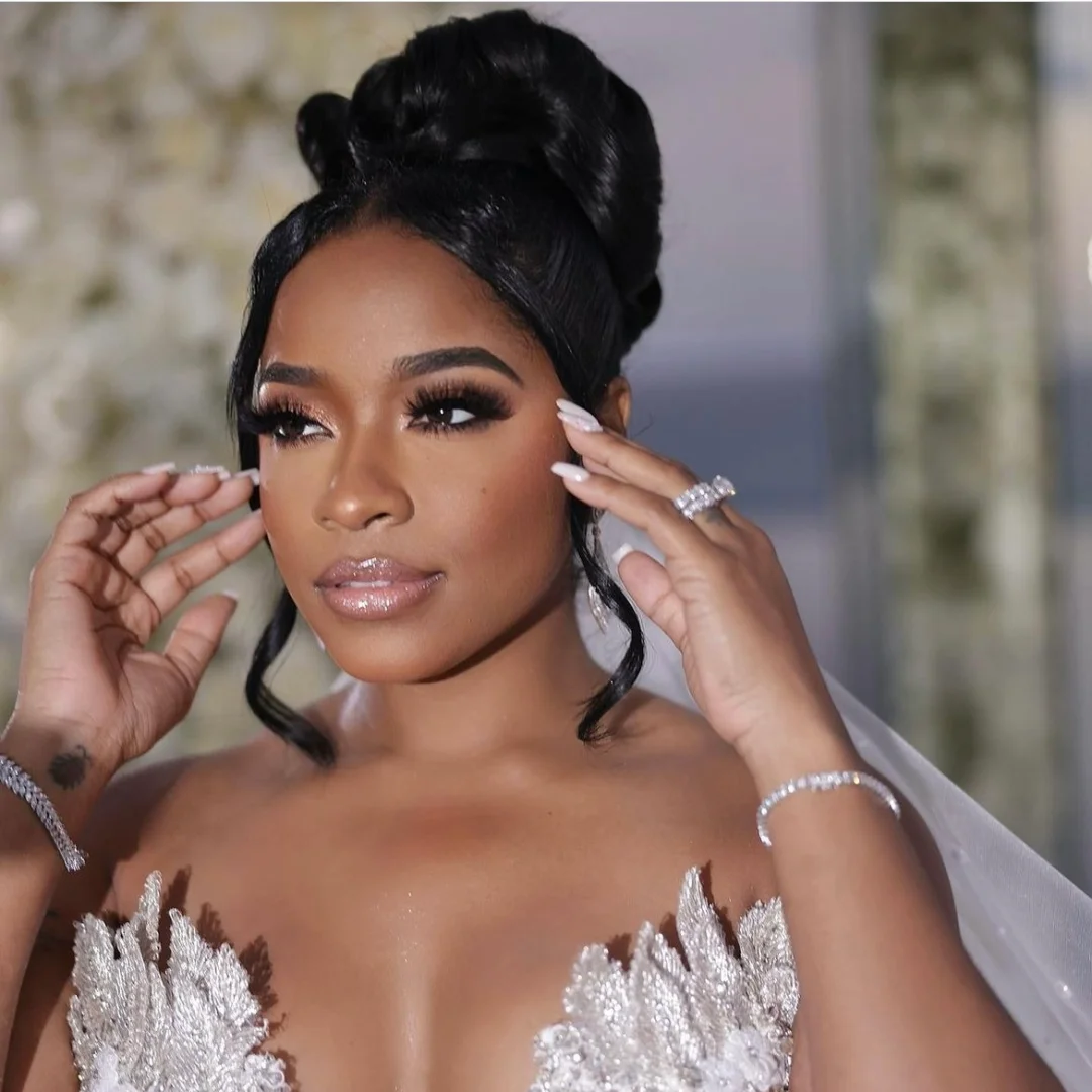 Beautiful bride with an elegant updo hairstyle, wearing a white wedding dress with intricate lace details, adjusting her hair and showing her rings, with a blurred floral background.